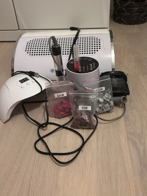 Nagelfilmaskin med tillbehör - Nagelfilmaskin med flera sliphuvuden och slipband i olika grovlekar (80#, 120#, 240#). Inkluderar elektrisk nagelfil, utsug, fotpedal och UV-lampa. Perfekt för nagelvård och manikyr hemma. Kan hitta nagel gelé fläckar på UV lampan och utsuget. Men annars alla alla saker i bra skick! Elfilen och utsuget är köpt på Amazon, elfilen kostade ungefär 450kr och utsuget ungefär 300kr. UV lampan är köpt på normal för 200kr.