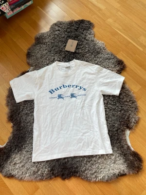 Vit Burberrys t-shirt med blått tryck - Snygg vit t-shirt från Burberrys med blått tryck på bröstet där det står 'Burberrys' och städerna London, Paris, New York. Klassisk rund hals och korta ärmar. Tillverkad i mjuk bomull för en skön känsla. Säljer den för att den inte kommit till användning.