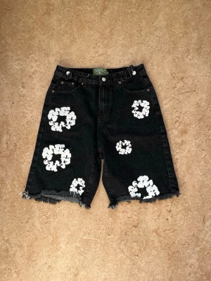 Svarta jeansshorts från Denim Tears - Svarta jeansshorts från Denim Tears med vita blommiga tryck och råa kanter. Klassisk femficksmodell med knappgylf och hög midja. Perfekta för dig som vill sticka ut med en unik streetwear-look.