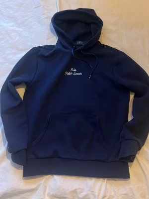 Mörkblå hoodie från Polo Ralph Lauren - Snygg mörkblå hoodie från Polo Ralph Lauren med broderad logga på bröstet. Klassisk huva med dragsko och stor magficka framtill. Tillverkad i mjukt material som känns skönt mot huden. Perfekt för en avslappnad och stilren look.