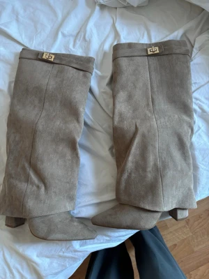 Beige höga mockastövlar med klack - Snygga beige stövlar i mockaimitation med spetsig tå och bred klack. Stövlarna har ett dekorativt guldfärgat spänne upptill ock en slouchy passform som ger en avslappnad men trendig look. Perfekta till höstens och vinterns outfits.