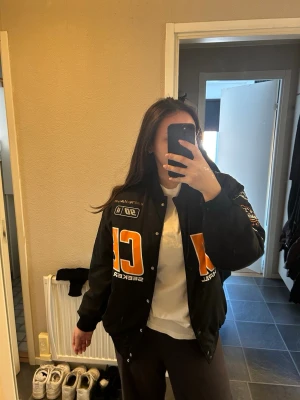 Svart bomberjacka med tryck - Snygg svart bomberjacka med stora orangea och vita tryck på bröstet och ärmen. Jackan har en oversized passform, tryckknappar och ribbade muddar. Perfekt för dig som gillar streetwear och sportig stil.