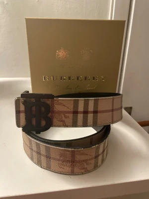 Burberry bälte med logga - Snyggt bälte från Burberry med klassiskt rutigt mönster i beige, brunt och rött. Bältet har en stor metallspänne med Burberrys ikoniska TB-logga. Perfekt accessoar för att lyfta vilken outfit som helst. Priset är inte hugget i sten!! Så kom med prisförslag!☺️😃😃
