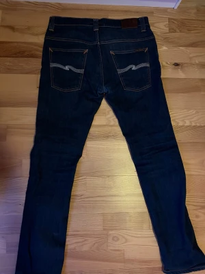 Mörkblå jeans från Nudie Jeans - Snygga mörkblå jeans från Nudie Jeans med klassisk femficksmodell och kontrastsömmar. Bakfickorna har Nudie Jeans karakteristiska broderade vågmönster. Jeansen är raka och smala i modellen.