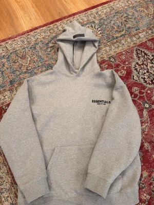 Grå Essentials hoodie - Säljer en grå hoodie från Essentials Fear of God med svart logga på bröstet och stor text på ryggen. Klassisk passform med huva och känguruficka framtill. Tillverkad i mjuk bomullsblandning som är perfekt för en avslappnad streetwear-look.