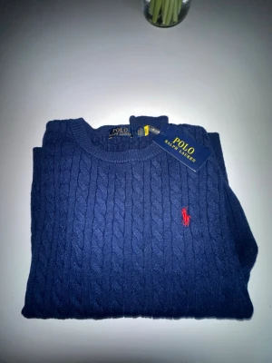 Blå kabelstickad tröja Polo Ralph Lauren - Mörkblå kabelstickad tröja från Polo Ralph Lauren med rund halsringning och röd broderad logga på bröstet. Klassisk och stilren design, perfekt för lager-på-lager. Tröjan har ribbade muddar och är tillverkad i mjukt material. Perfekt passad till Storleken S, den kan se stor ut när den ligger ner på borden men som du ser på bilden så passar den perfekt när man har den på kroppen. Aldrig använd förutom när jag tog bilden och hade på mig den i 20 sekunder.