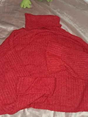 Röd ribbad polotröja stickad - Mysig röd stickad polotröja med ribbad struktur. Tröjan har långa ärmar och en hög polokrage som ger extra värme. Perfekt för kyliga dagar och enkel att styla till olika outfits.