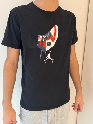 Svart Nike Air Jordan t-shirt - Svart t-shirt från Nike med stor Air Jordan-sneaker och Jumpman-logga tryckt på bröstet. Röd Nike-logga och swoosh på ryggen. Klassisk passform och rund halsringning. Storleken stämmer ej som är på bilden, den passar xs-s