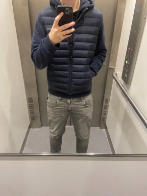 Mörkblå dunväst från J.Lindeberg - Snygg mörkblå dunväst från J.Lindeberg med diskret logga på bröstet. Västen har en quiltad design, hög krage och dragkedja framtill. Perfekt att slänga över en hoodie för en stilren look. Materialet är lätt och värmande. Perfekt nu när våren börjar komma!