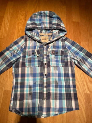 Rutig blå skjorta med huva Hollister - Säljer en rutig skjorta från Hollister i blå, vit och turkos med huva och knappar framtill. Skjortan har två bröstfickor med knapp och en avslappnad passform. Perfekt för dig som gillar en chill och casual stil.