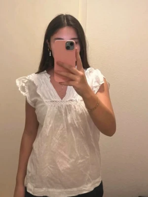 Vit blus från gina tricot  - Söt vit blus med v-ringning, volangdetaljer vid axlarna och spets längs halsringningen. Blusen är luftig och har en romantisk känsla, perfekt till sommaren. Tillverkad i ett lätt tyg som känns skönt mot huden.