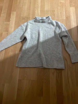 Ljusgrå stickad tröja med hög krage - En enkel och stilren ljusgrå stickad tröja med högre krage och långa ärmar. Tröjan har en rak passform och ribbstickade muddar vid ärmslut och nederkant. Perfekt för lager på lager och passar till det mesta.