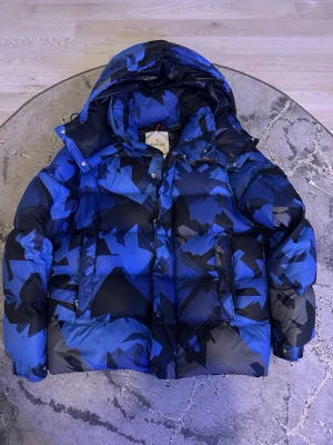 Blå och svart dunjacka från Moncler - Säljer en fet blå och svart dunjacka från Moncler med coolt geometriskt mönster. Jackan har stor huva, dragkedja och tryckknappar framtill samt två stora fickor. Perfekt för kalla vinterdagar och riktigt snygg streetstil.