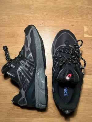 Asics Gel NYC - Säljer ett par snygga Asics sneakers i svart och grått med coola detaljer och mönstrade skosnören. Skorna har en robust sula och meshpartier för extra komfort. Perfekta för dig som gillar sportig stil och vill ha något som funkar till både jeans och träningskläder. I storlek 42,5