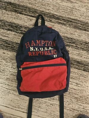 Blå och röd ryggsäck Hampton Republic - Snygg ryggsäck från Hampton Republic i marinblått tyg med röd frontficka och röda axelband. Stor ficka framtill med dragkedja och tryck i vitt och rött på framsidan. Perfekt för skolan eller utflykter, klassisk och sportig design.