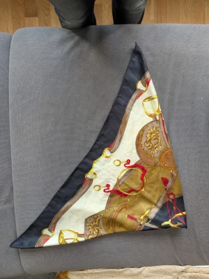 Mönstrad sjal med kedjedetaljer - Säljer en snygg sjal med mönster av kedjor och rep i guld, rött och brunt på en vit bakgrund. Sjalen har en svart kant, perfekt att styla runt halsen eller i håret. Materialet känns glansigt och liknar siden.