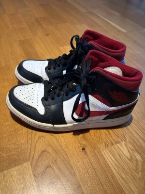 Nike Air Jordan 1 Mid svart/vit/röd - Säljer ett par Nike Air Jordan 1 Mid sneakers i svart, vitt och rött. Skorna har klassisk hög siluett, svart läderdetaljer, vit bas och röda accenter samt röd insida. Ikonisk vinge-logga på sidan och svart snörning. Lite slitna vid tårna se bild 