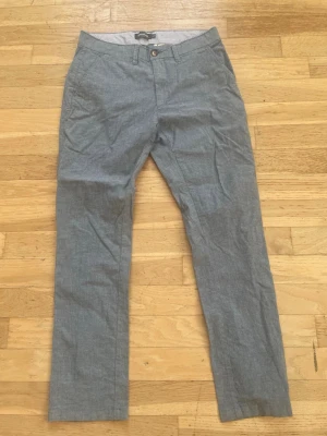 Grå linnebyxor från Dressmann - Snygga grå linnebyxor från Dressmann i slim fit-modell. Byxorna har klassisk knappgylf, bälteshällor och diskreta sidofickor. Perfekta för en avslappnad men stilren look. Materialet är lätt och andas, vilket gör dem sköna att bära.