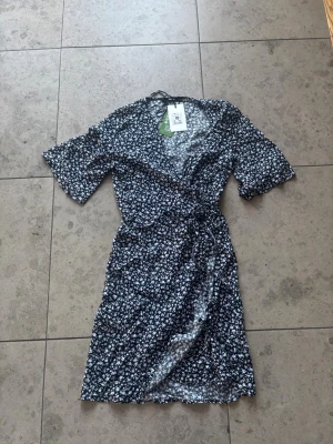 Blommig omlottklänning från Bik Bok - Supersöt omlottklänning från Bik Bok i marinblått med vitt blommönster. Klänningen har kort ärm, knyt i midjan och v-ringad hals. Perfekt för en fräsch och somrig look.