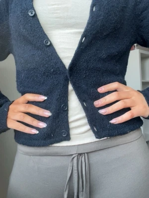 Mörkblå fluffig kofta med knappar - Mysig mörkblå kofta med rund hals och knäppning framtill. Koftan är fluffig och har långa ärmar, perfekt för lager på lager. Enkel och stilren design som passar till det mesta. Materialet ser ut att vara en mjuk ull- eller mohairblandning.