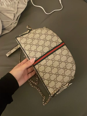 Gucci beige axelväska med kedja - Säljer en beige axelväska från Gucci med klassiskt GG-mönster och en röd-grön rand på framsidan. Väskan har guldfärgad kedjerem och detaljer i läder. Perfekt storlek för det viktigaste och riktigt snygg till alla outfits.