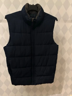 Mörkblå dunväst från GAP - Snygg mörkblå dunväst med hög krage och quiltad design. Västen har dragkedja framtill och en stilren, enkel look som passar perfekt till lager-på-lager. Fodrad insida för extra värme och komfort. Perfekt för kyliga dagar när du vill hålla stilen.