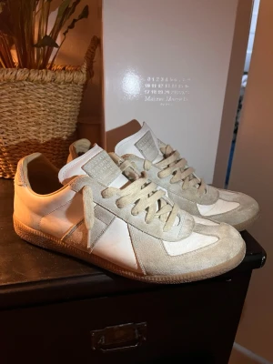 Maison Margiela  - Maison Margiela Replica sneakers i beige mocka och vitt läder. Klassisk low-top modell med beige snörning och gummisula. Ikonisk design med diskreta detaljer och stilren look. Perfekta för dig som gillar minimalistisk och trendig stil. Gör noggrant rent dom innan inlämning! Storlek 45. BOX OCH SHOEBAGS INGÅR! 