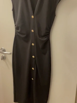 Svart klänning med guldknappar - Stilren svart klänning med v-ringning och dekorativa guldknappar framtill. Klänningen har en smickrande passform och är ärmlös, perfekt för dig som gillar en klassisk look med en twist.