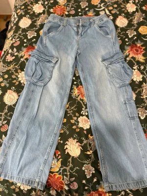 Ljusblå low waist cargobyxor - Säljer ett par ljusblå cargobyxor i denim med bred passform och flera stora fickor på sidorna. Byxorna har resår i midjan och knappstängning.            . Perfekta för en avslappnad och trendig look.         Innerbenslängd: 72 cm höft :70 cm( men väldigt stretchig)