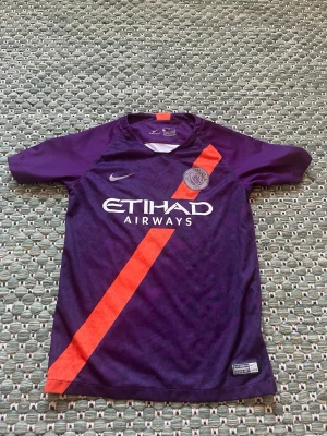 Lila Manchester City matchtröja Nike - Snygg lila Manchester City matchtröja från Nike med orange diagonal rand och klubbmärke på bröstet. Tröjan har korta ärmar, rund hals och är tillverkad i ett lätt, andningsbart funktionsmaterial. Perfekt för fotbollsträning eller att bära till vardags.
