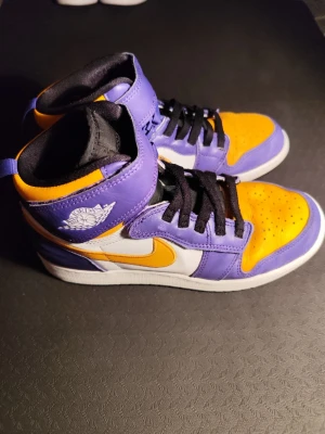 Air Jordan 1 High Lakers färg - Säljer ett par Air Jordan 1 High sneakers i Lakers-inspirerad färgkombination med lila, gult och vitt. Skorna har klassisk hög siluett, svart snörning och vit yttersula. Tillverkade i läder med ikoniska Air Jordan-loggan på sidan. Perfekta för dig som gillar streetwear och basketstil.