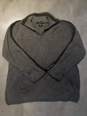 Grå stickad tröja från Calvin Klein - Mörkgrå stickad tröja från Calvin Klein Jeans med halv dragkedja och ribbad struktur. Tröjan har svarta detaljer längs ärmarna och diskret logga på ena ärmen. Skickas inom 24 timmar🚀📦