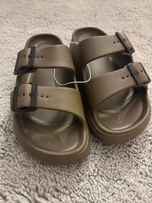 Bruna sandaler med dubbla spännen - Snygga bruna sandaler med två breda remmar och justerbara svarta spännen. Tillverkade i ett lätt och tåligt plastmaterial med formad fotbädd för extra komfort. Perfekta för sommarens alla äventyr. Dem är även ny köpta utan prislapp! pris kan diskuteras 