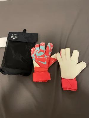 Nike målvaktshandskar röd/blå - Säljer ett par Nike GK Grip3 målvaktshandskar i rött och ljusblått med vita handflator. Handskarna har breda kardborreband runt handleden för extra stöd och grepp. Perfekta för fotbollsmatcher och träning. Kommer med original svart förvaringspåse., ALDRIG ANVÄNT!!!!