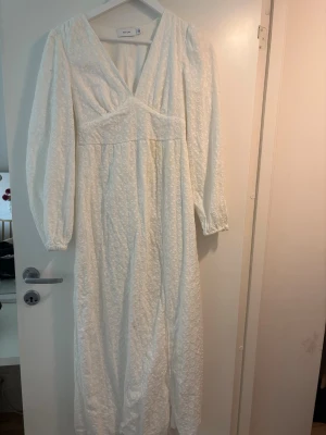 Vit långklänning från Vila - Långklänning i vitt från Vila med v-ringning och långa ärmar. Klänningen har broderade blommor över hela tyget och är tillverkad i bomull. Perfekt för sommarens alla tillfällen och har en romantisk, luftig känsla. Klänningen har dock en fläck på sig, som man ska kunna få bort i tvätten. 
