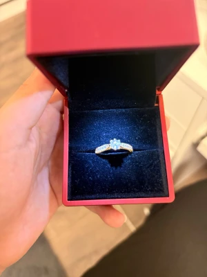Elegant silverring med gnistrande stenar - Säljer en elegant silverfärgad ring med en större klar sten i mitten och mindre stenar längs bandet. Ringen har en klassisk och tidlös design som verkligen fångar ljuset. Perfekt för dig som gillar stilrena smycken med lite extra bling.