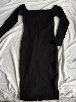 Svart långärmad offshoulder klänning (S) - Stilren svart långklänning med lång ärm och offshoulder-design. Klänningen har en figurnära passform och är tillverkad i ett stretchigt material som ger en snygg siluett. Perfekt för dig som gillar minimalistisk och elegant stil.