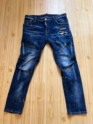 Mörkblå distressed jeans Dsquared2 - Säljer ett par mörkblå jeans från Dsquared2 med slitningar, färgstänk och patchdetaljer. Jeansen har smal passform, klassisk femficksdesign och orange kontrastsömmar. Perfekta för dig som gillar en edgy streetstil.
