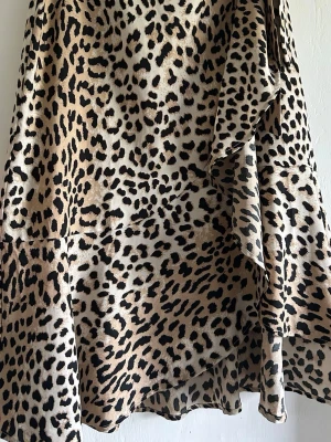 Leopardmönstrad omlottkjol med knyt - Snygg omlottkjol med leopardmönster i beige och svart. Kjolen har en feminin volangkant och knyt i midjan för en justerbar passform. Perfekt statement-plagg för dig som vill sticka ut. Tråden till knappen är lite lös 
