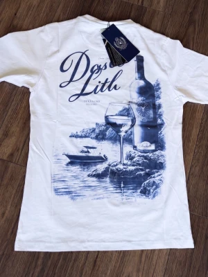 Vit t-shirt med marinblått tryck - Säljer en vit t-shirt från Leon Marcel med ett stort marinblått tryck på ryggen som visar en vinflaska, ett vinglas, en båt och klippor vid vattnet. T-shirten har rund hals och korta ärmar. Perfekt för dig som gillar stilrena motiv med sommarkänsla.