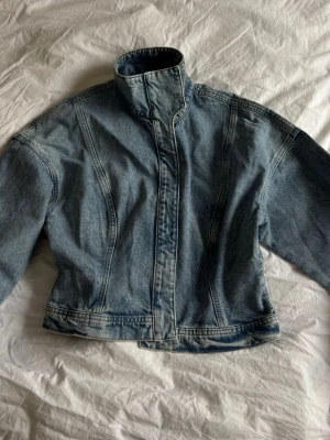 Ljusblå croppad jeansjacka - Snygg ljusblå jeansjacka med hög krage och markerad midja. Jackan har en croppad passform, dolda knappar framtill och sömdetaljer som ger en cool vintagekänsla. Perfekt att slänga över en hoodie eller t-shirt för en avslappnad look.