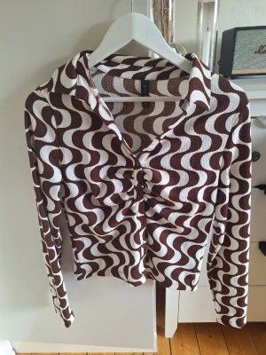 Brunvitrandig retroblus från new yorker - Säljer en brun och vit blus från new yorker med vågigt retroinspirerat mönster. Blusen har lång ärm, krage och knappar framtill med snygg rynkning vid bröstet. Materialet är stretchigt och strukturerat för en cool look.