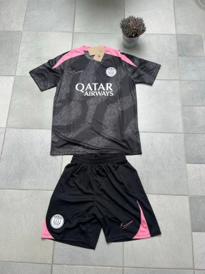 PSG svart och rosa matchställ Nike  - Snyggt PSG matchställ från Nike i svart med rosa detaljer. Tröjan har korta ärmar, tryckt PSG-logga och Qatar Airways på bröstet. Shortsen är svarta med rosa inslag och PSG-märke. Perfekt för fotbollsträning eller streetstyle. Storlek S