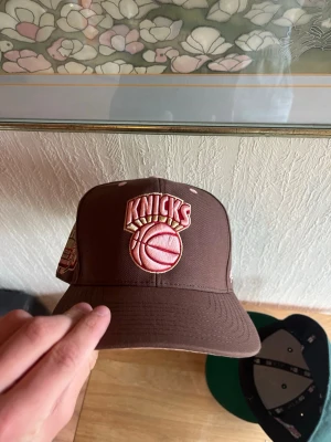 Brun Knicks keps NBA - Säljer en brun snapback-keps med New York Knicks broderad logga framtill och NBA Hardwood Classics-märke bak. Kepsen har platt skärm och är gjord i bomullstyg. Perfekt för dig som gillar basket och streetwear. 61 cm i storlek