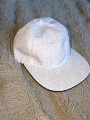 Vit keps från Fendi med FF-mönster - Säljer en stilren vit keps från Fendi med det klassiska FF-mönstret ton-i-ton över hela kepsen. Kepsen har böjd skärm och justerbar rem bak för perfekt passform. Materialet är en strukturerad bomullsblandning som känns både lyxig och bekväm.