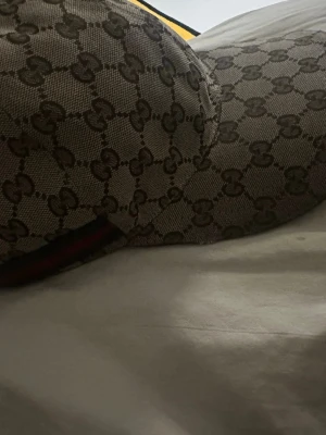 Gucci keps med GG-mönster - Säljer en snygg keps från Gucci med klassiskt GG-monogram i beige och brunt. Kepsen har böjd skärm och justerbar rem baktill. Materialet är canvas med detaljer i svart och röd rand. Perfekt för dig som vill sticka ut med en lyxig accessoar.   Köpt på gucci butiken i Stockholm City 100% äkta, pris kan diskuteras vid snabb affär.