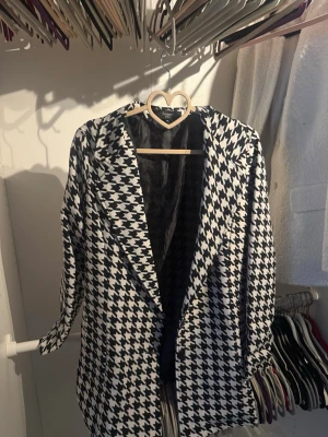 Houndstooth kappa från Sisters Point - Snygg svartvit kappa från Sisters Point i klassiskt hundtandsmönster. Oversized passform med breda slag och helfodrad insida. Perfekt statement-plagg för dig som vill sticka ut med din stil. Passar till lager på lager och ger en trendig look.
