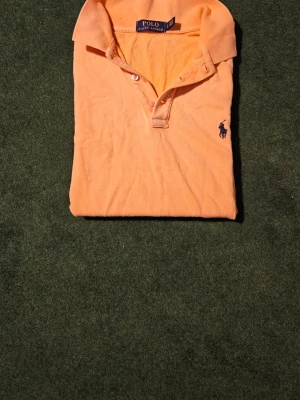 Orange pikétröja från Polo Ralph Lauren - Klassisk orange pikétröja från Polo Ralph Lauren med blå logga broderad på bröstet. Tröjan har krage, knappar framtill och är tillverkad i mjuk bomull. Perfekt för en avslappnad men ändå stilren look. Lite blekt men användbar.