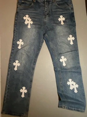 Levi's X Chrome Hearts jeans blåa - Unika blå Levi's X Chrome Hearts jeans med raka ben och flera vita korspatches sydda över hela byxorna. Klassisk femficksmodell i slitstark denim, perfekt för dig som vill sticka ut med en streetwear-inspirerad.