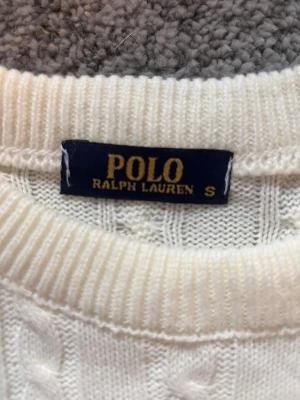 Vit kabelstickad tröja Polo Ralph Lauren - Vit kabelstickad tröja från Polo Ralph Lauren med klassisk svart broderad logga på bröstet. Tröjan har rund halsringning och är gjord i mjukt stickat material. Perfekt för en stilren och tidlös look. Priset går att diskutera. Ask i bild medföljer ej